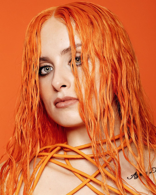 A close-up portrait of a woman with striking orange hair and intense amber eyes, against a warm orange background. orange wires are draped around her shoulders.