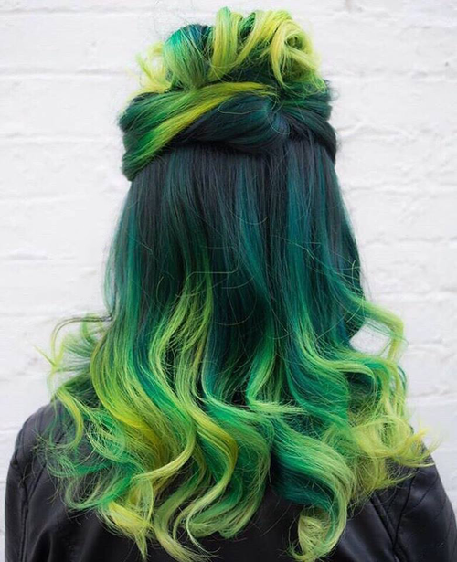 A person seen from behind showcasing vibrant hair dyed in shades of green and yellow, styled with a top bun and wavy lengths against a white brick wall.