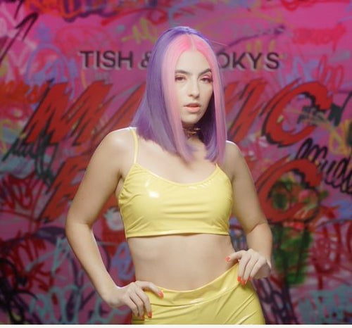 A woman with pink an purple hair, posing in front of a colorful graffiti-covered wall that say Manic Panic
