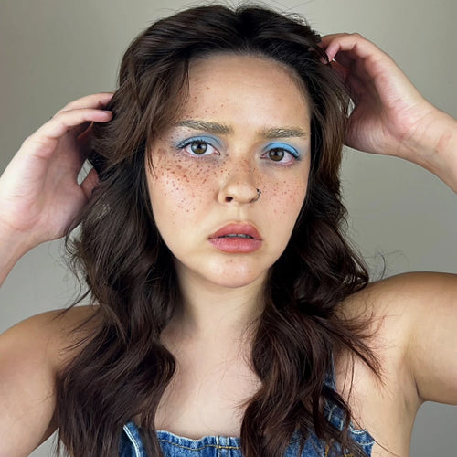 Woman with freckles and blue eyeshadow adjusting her hair against a neutral background
