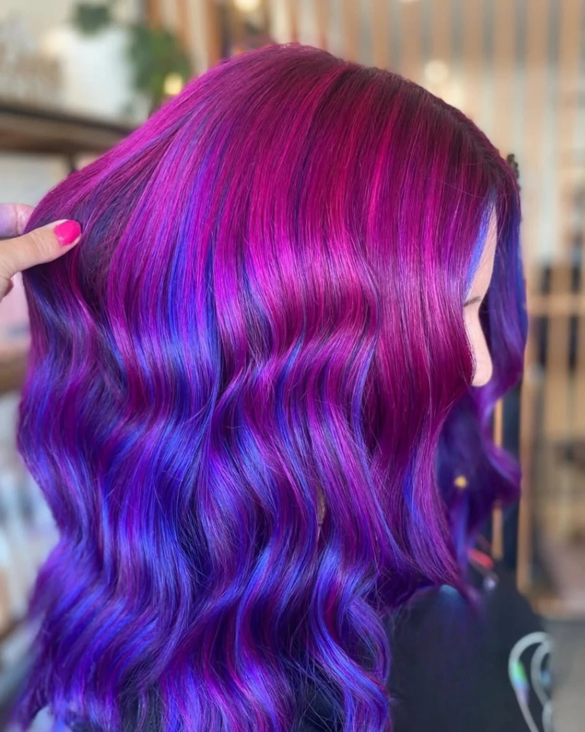 A close-up of a person with long, wavy hair styled in vibrant magenta and purple tones, with a hand lifting a section of the hair to show texture and color. The background is a softly blurred indoor setting.
