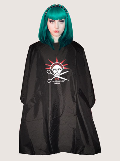 Woman with green hair wearing a salon cape with a prominent skull and cross bone logo with head spikes similar to the statue of liberty